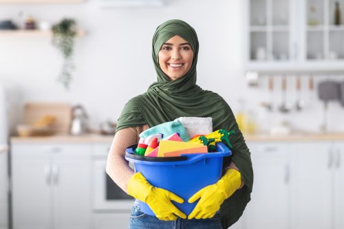 Smiling muslim female house-keeper cleaning apartment, holding blue bucket with cleaning tools and rags, standing in modern kitchen interior, copy space. Cheerful young woman housekeeping on weekend